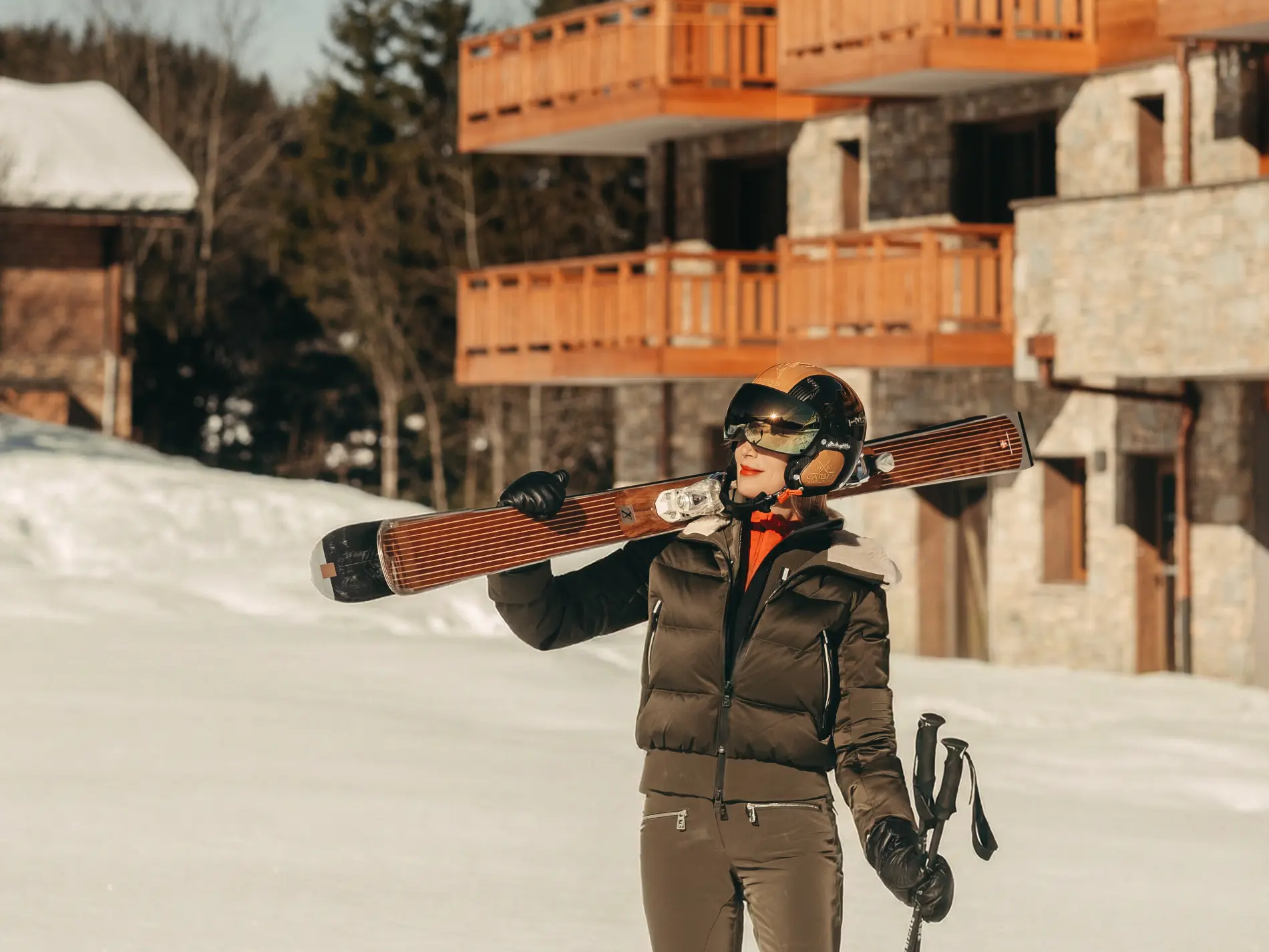 Femme en tenue de ski kaki tenant des skis en bois sur l'épaule devant l'hôtel Bârma à Courchevel La Tania sous la neige