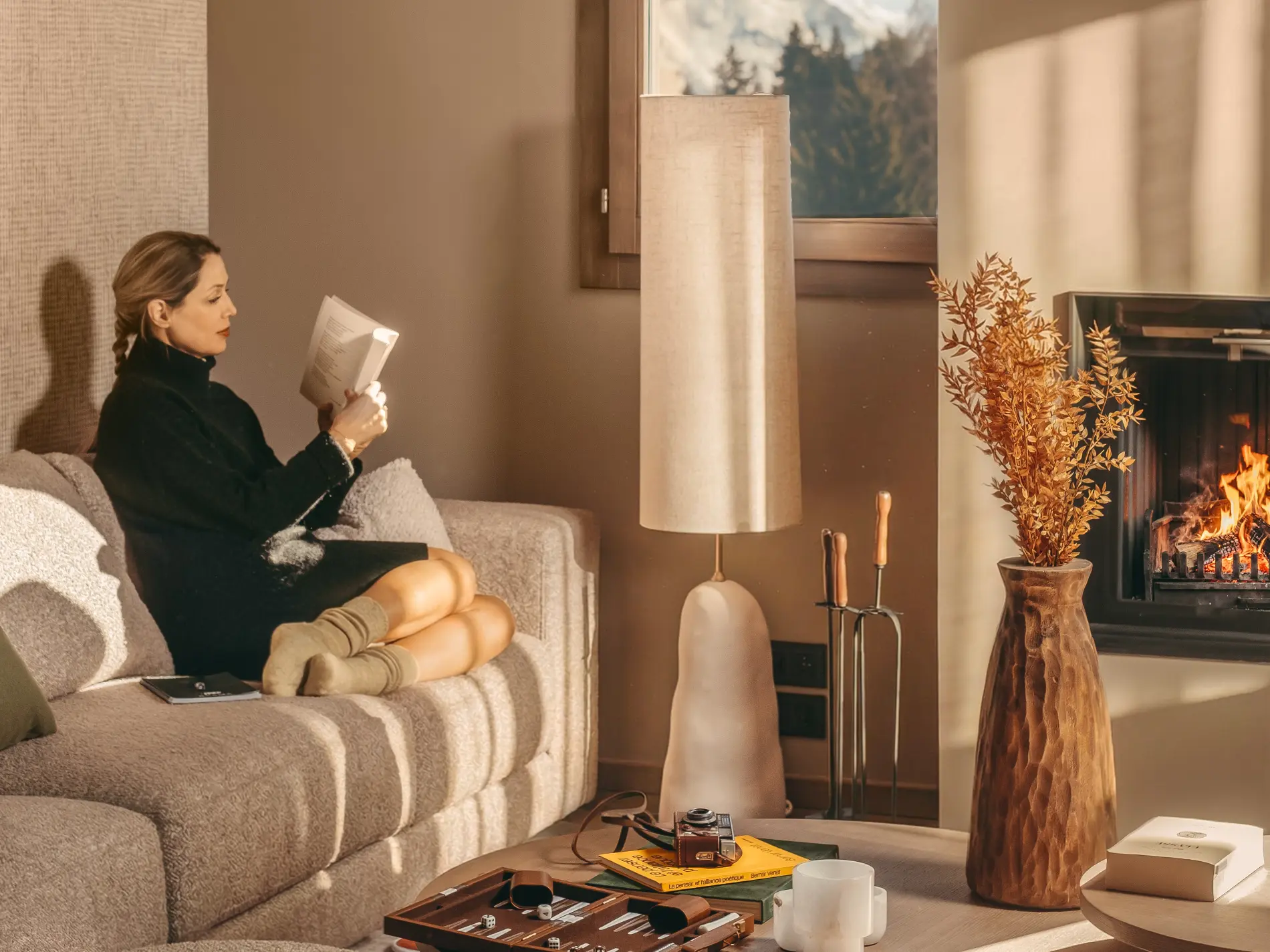 Femme lisant un livre sur un canapé près d'une cheminée allumée dans le salon d'une suite de l'hôtel Bârma à Courchevel La Tania, vue sur les sommets enneigés