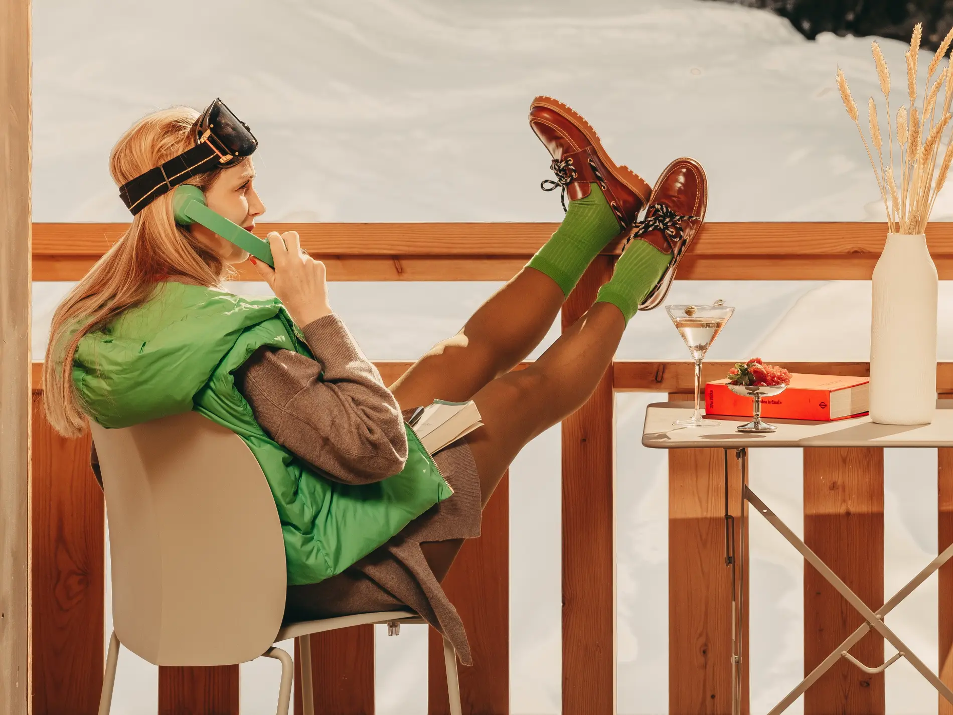 Jeune femme en doudoune verte parlant au téléphone vintage, pieds sur la table, martini à portée de main sur la terrasse enneigée de l'hôtel Bârma à Courchevel La Tania