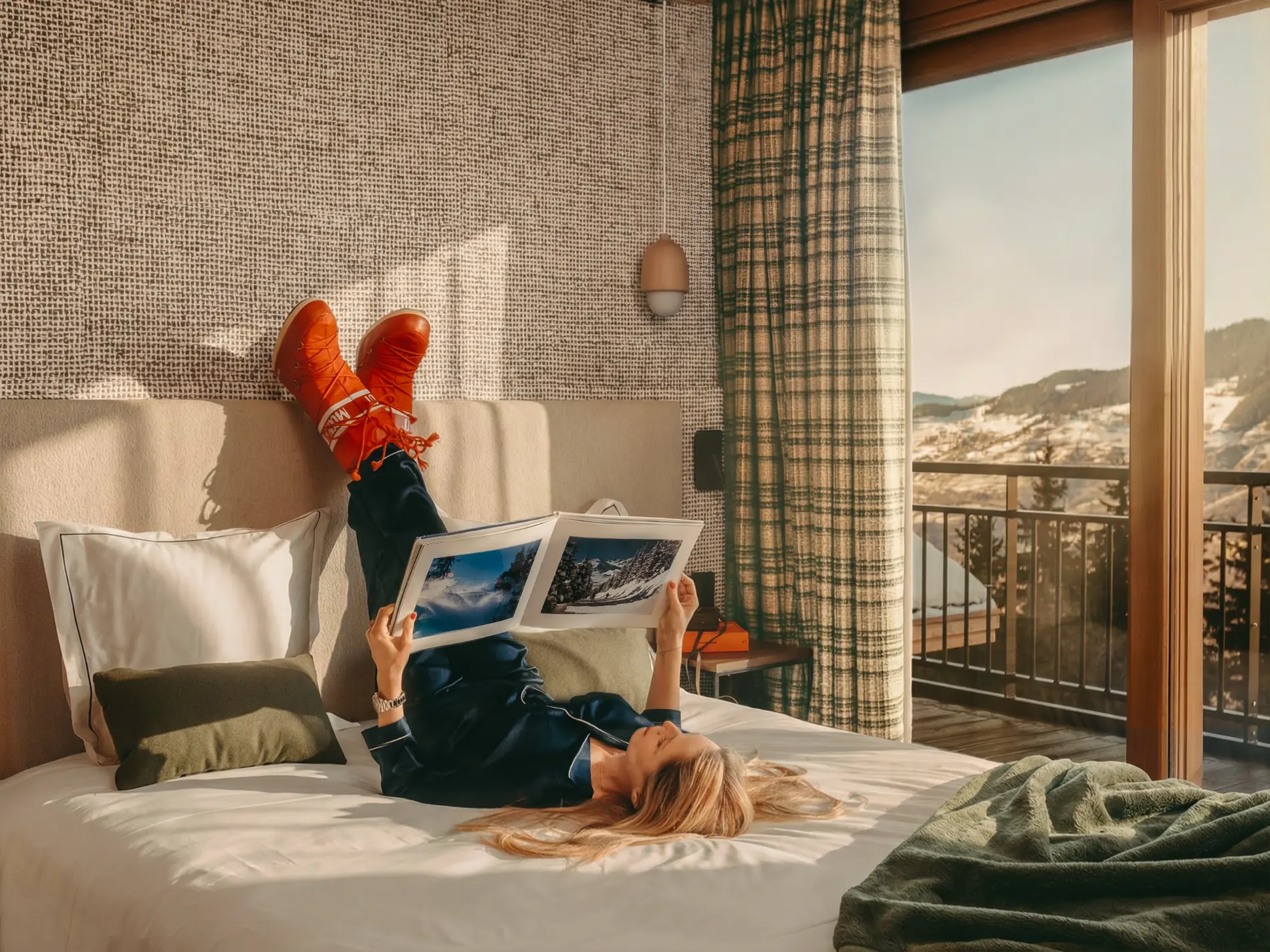 Femme en pyjama bleu et Moon Boots oranges allongée sur un lit blanc, lisant un magazine de ski dans une chambre de l'hôtel Bârma à Courchevel La Tania avec panorama sur les Alpes